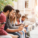 Studentinnen und Studentinnen sitzen zusammen vor ihrer Hochschule und schauen gemeinsam auf Laptops und Smartphones